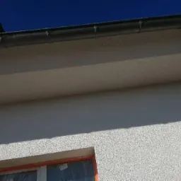 Fragment elewacji budynku z widocznym orynnowaniem i oknem zabezpieczonym folią, świeżo po nałożeniu tynku strukturalnego.