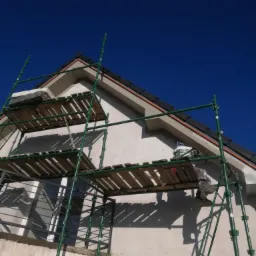 Praca na wysokości: ocieplanie elewacji domu jednorodzinnego z użyciem rusztowania, widoczna puszka z zaprawą i fragment elewacji po nałożeniu warstwy ociepleniowej.