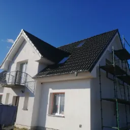 Nowo otynkowany dom jednorodzinny z grafitową dachówką i rusztowaniem z boku, widok z dołu na tle błękitnego nieba.