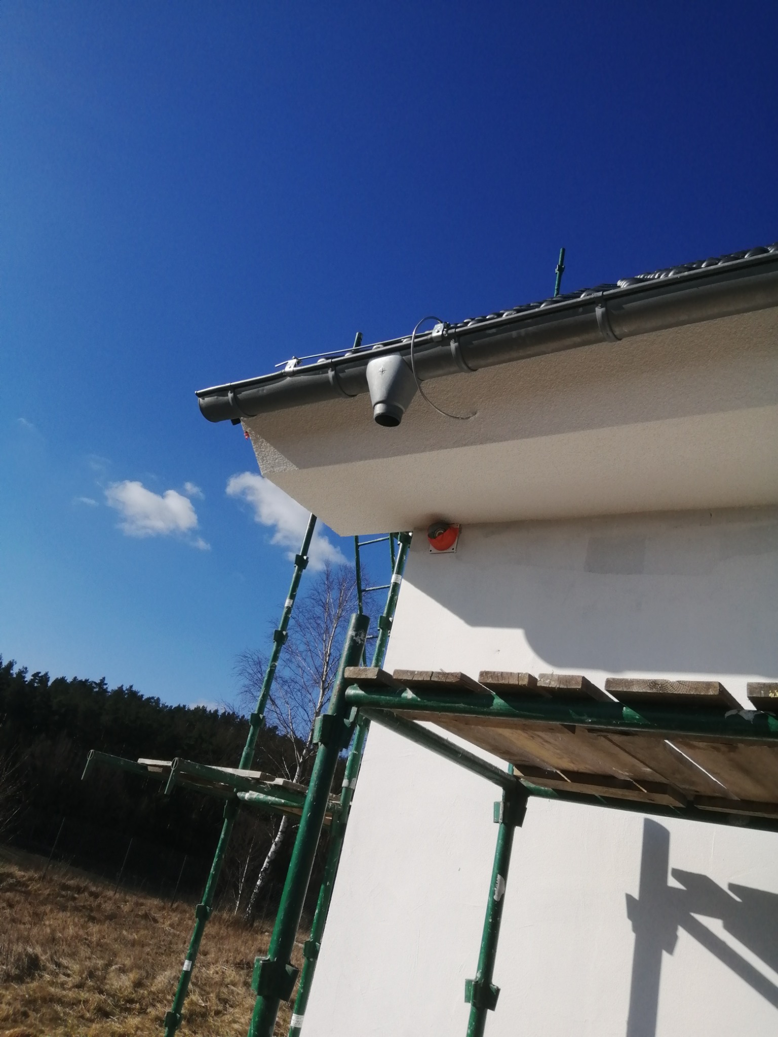 Fragment ocieplonej ściany budynku z zamontowaną rynną, widoczny fragment rusztowania na tle błękitnego nieba i lasu.