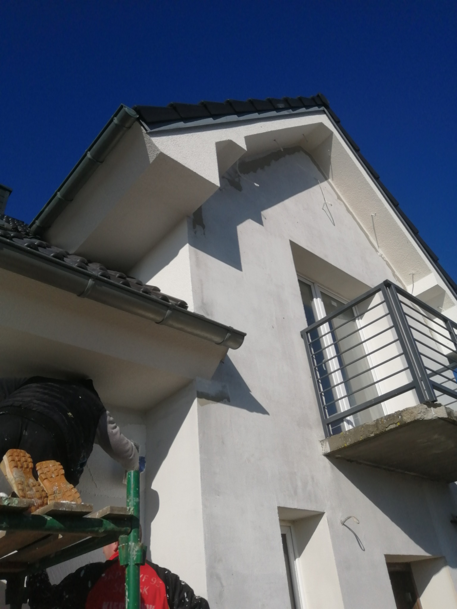 Praca na rusztowaniu przy białej elewacji domu z balkonem, widoczne buty robotnika i fragment czerwonej kurtki, błękitne niebo w tle.