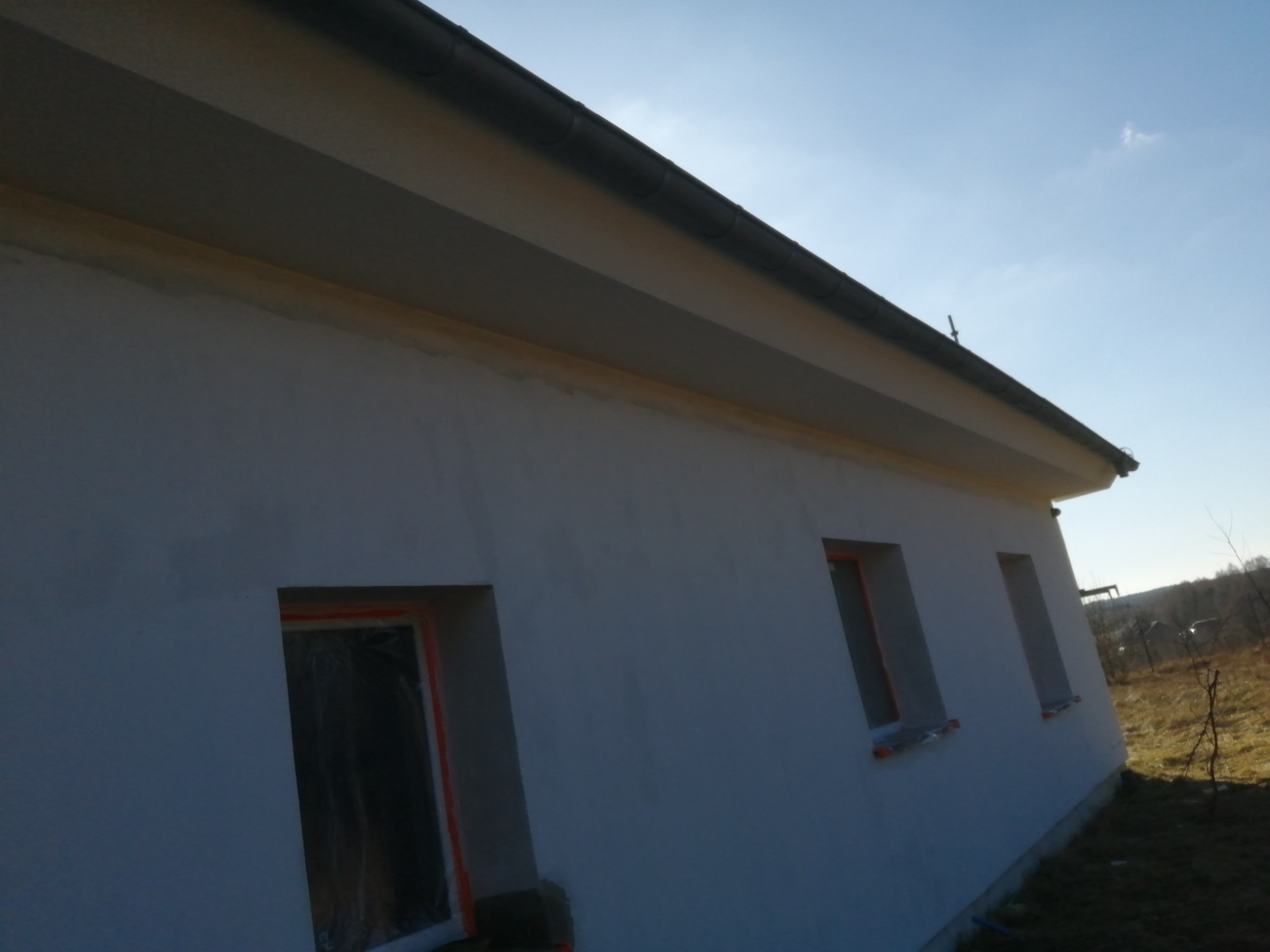 Świeżo otynkowana elewacja domu jednorodzinnego z zabezpieczonymi oknami folią malarską, widok z dołu na jasnym tle nieba.