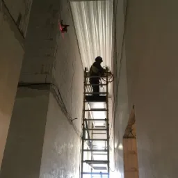 Elektryk na drabinie w surowym wnętrzu w Siedlcach, instalujący przewody, widoczny laserowy niwelator na ścianie z bloczków.