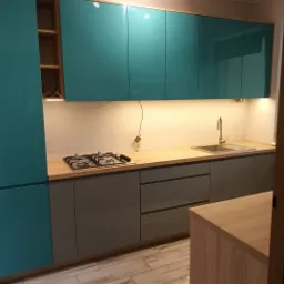 Nowoczesna kuchnia z turkusowymi szafkami górnymi z połyskiem i szarymi szafkami dolnymi, blatem drewnopodobnym oraz płytą gazową, widok na fragment podłogi z paneli imitujących drewno.