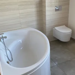 Nowoczesna, biała wanna narożna z chromowaną baterią, podwieszana toaleta i beżowe płytki imitujące drewno w wykończonej łazience.