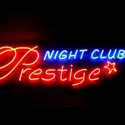 Neonowy szyld 'Prestige NIGHT CLUB' z czerwoną, ozdobną czcionką 'Prestige' i niebieskim napisem 'NIGHT CLUB' oraz czerwoną gwiazdką na czarnym tle.