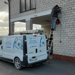 Instalator na drabinie montuje przewody na ścianie budynku, na dachu samochodu firmy IPDSystem drabina, na samochodzie reklama systemów alarmowych, kontroli dostępu i telewizji przemysłowej.