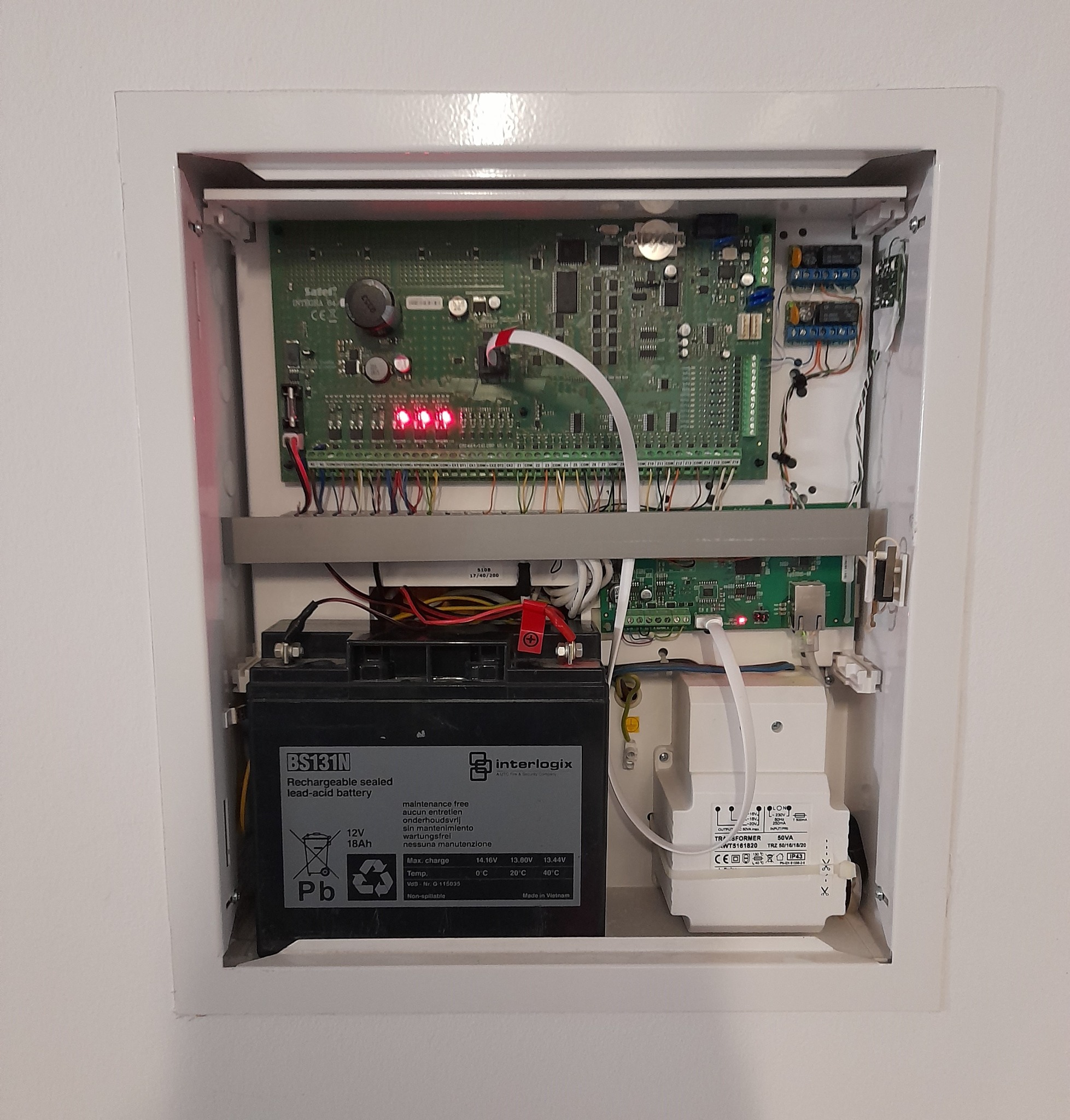Otwarta szafka z centralą alarmową Satel Integra 64, akumulatorem 12V 18Ah Interlogix i transformatorem TRZ 50/16/18/20, widoczne diody LED i okablowanie.