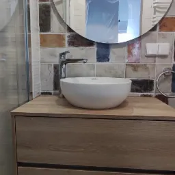 Umywalka nablatowa w kształcie misy na szafce z drewnopodobnym fornirem, nad nią okrągłe lustro i dekoracyjne płytki ścienne w odcieniach brązu i beżu, na podłodze wzorzyste płytki.