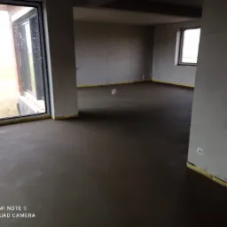 Świeżo wylana, jeszcze wilgotna, ciemna posadzka w surowym wnętrzu z dużymi oknami i żółtą taśmą zabezpieczającą przy ścianach.