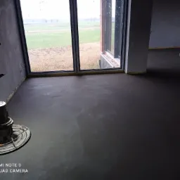 Świeżo wylana, szara posadzka w surowym wnętrzu z widokiem na pole przez duże okno; widoczna maszyna do zacierania posadzek.