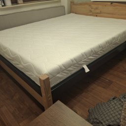 OLTECH Adam Oleksy - Drewniane łóżko z materacem, z widocznym stelażem i zagłówkiem. Na podłodze panele. Częściowo widoczna szafka nocna i kanapa. Stonowana kolorystyka.