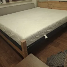 Drewniane łóżko z materacem, z widocznym stelażem i zagłówkiem. Na podłodze panele. Częściowo widoczna szafka nocna i kanapa. Stonowana kolorystyka.