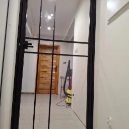 Czarna, metalowa ścianka działowa z szybami w korytarzu. Widoczna drewniana, ażurowa  ściana w tle. Minimalistyczny design,  jasne wnętrze.