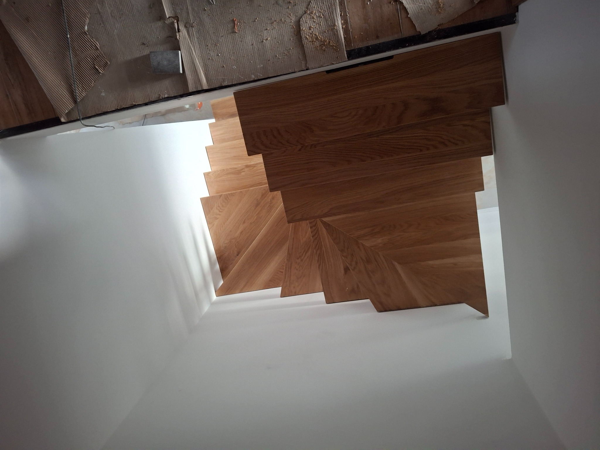 Dębowe schody z drewnianymi stopniami, widok z góry na białe ściany klatki schodowej. Fragment kartonowego zabezpieczenia widoczny u góry.