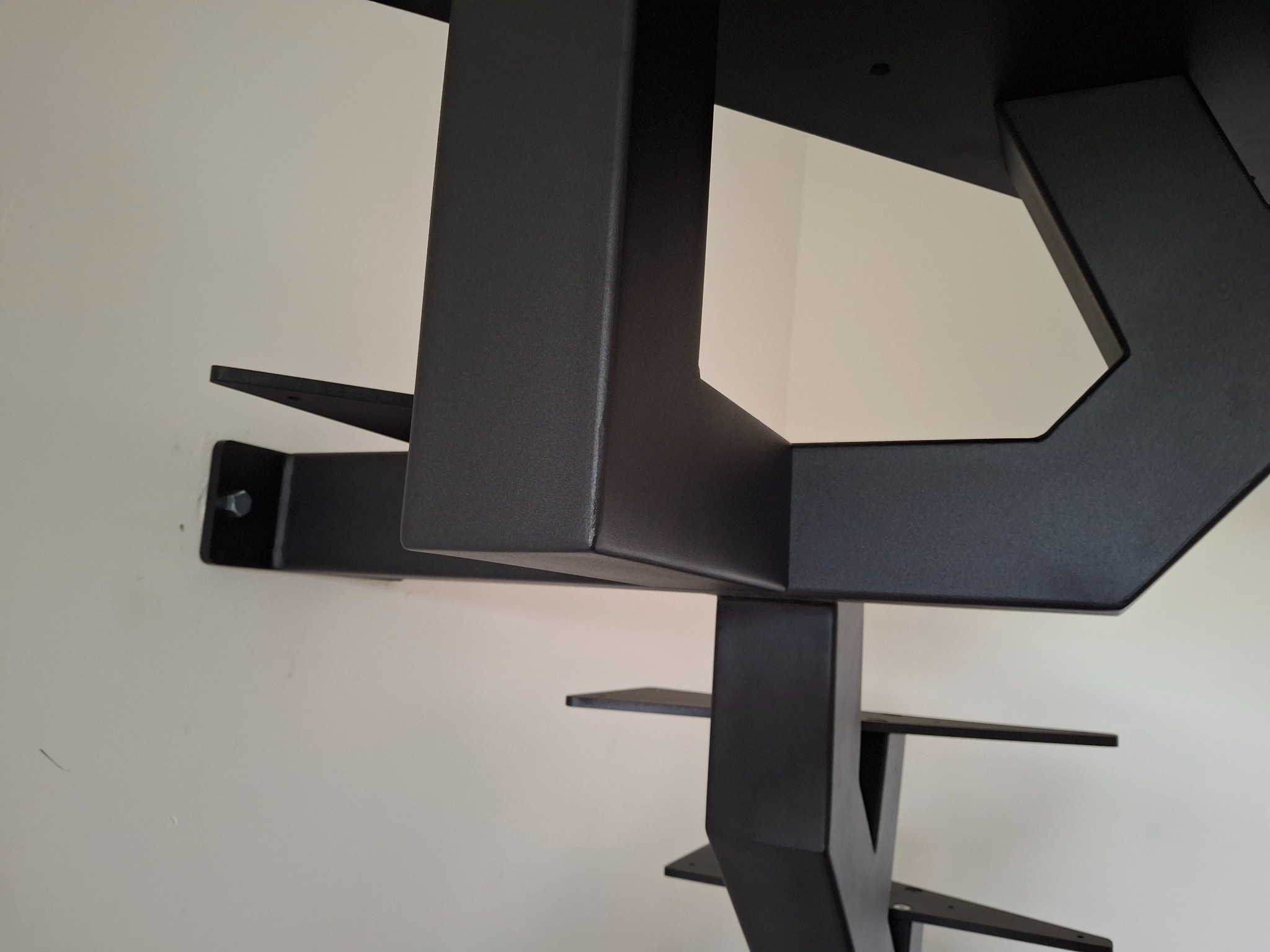 Nowoczesne, czarne schody metalowe o geometrycznej konstrukcji, przymocowane do jasnej ściany, widoczne z dołu pod kątem, z wyraźnymi detalami mocowań.