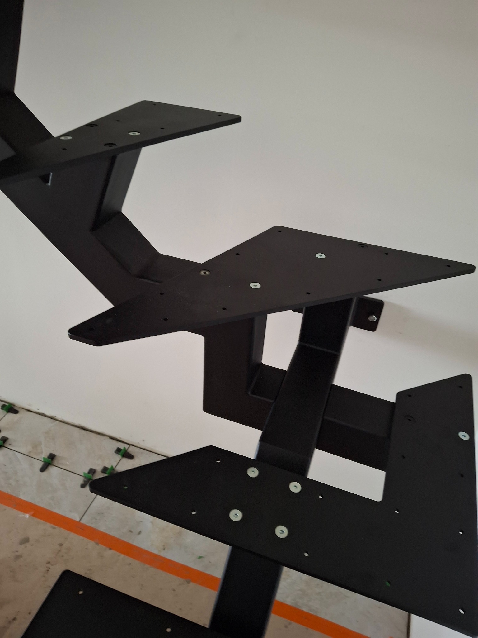 Czarne, metalowe elementy konstrukcji schodów z widocznymi śrubami montażowymi, zamontowane na ścianie. Podłoga z jasnych płytek z krzyżykami dystansowymi.