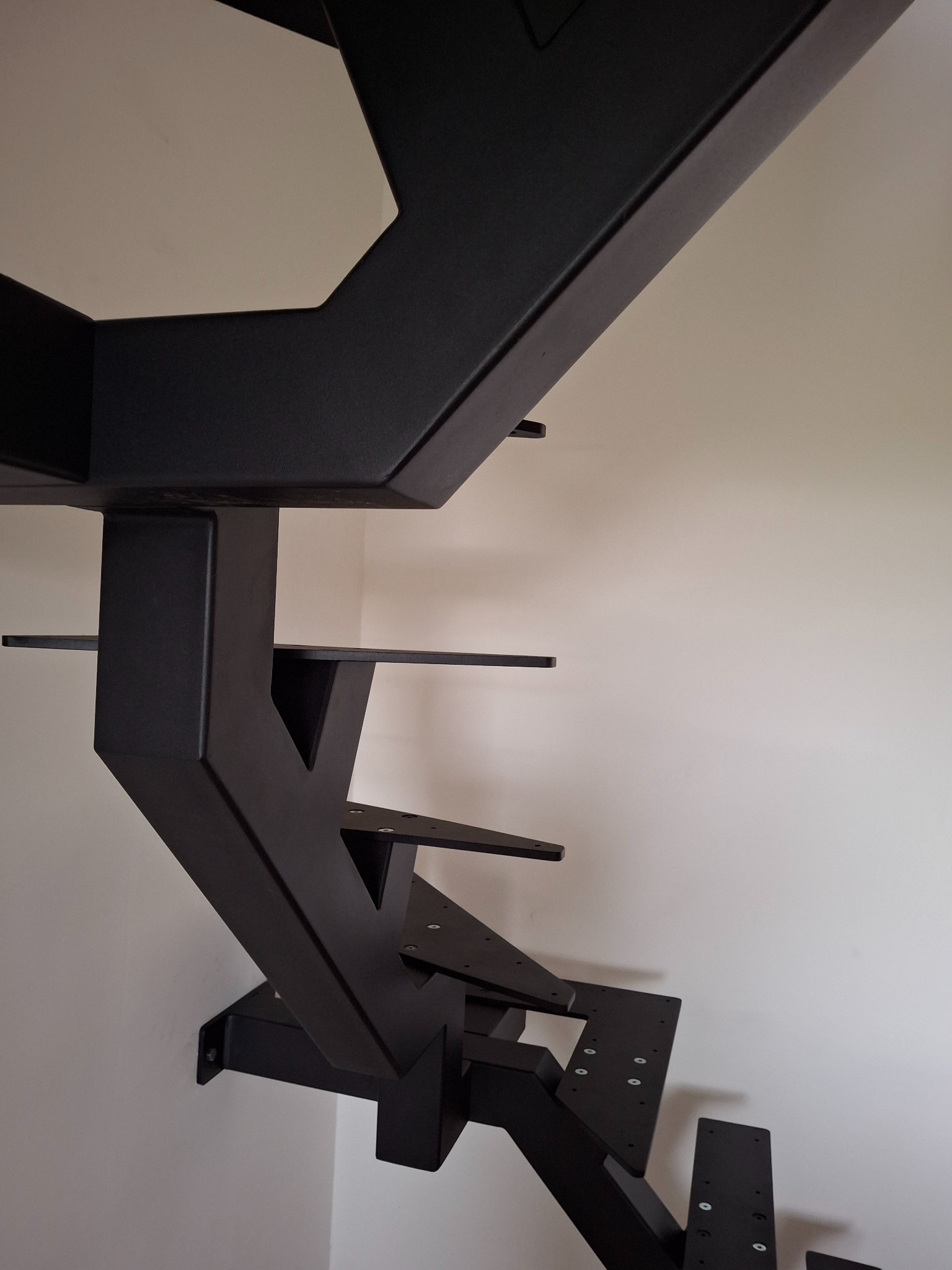 Nowoczesne, czarne schody metalowe przy ścianie, widok z dołu, ukazujący konstrukcję wspornikową i minimalistyczny design. Mocowania do ściany widoczne.