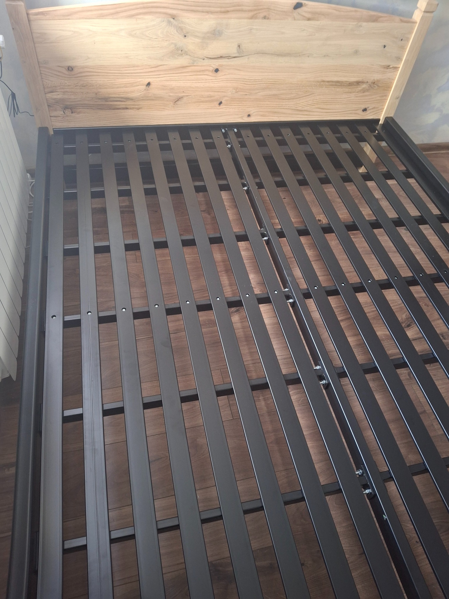 Rama łóżka z metalowymi szczebelkami i drewnianym zagłówkiem, widoczna z góry pod kątem, z elementami montażowymi.