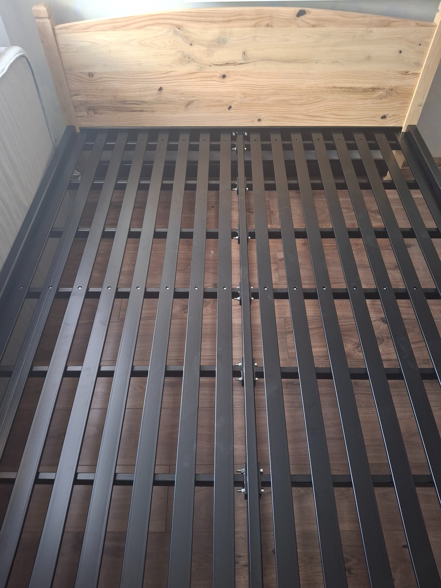 Metalowa rama łóżka z drewnianym zagłówkiem, widok z góry. Konstrukcja z ciemnych, metalowych listew na drewnianej podłodze. Surowy styl.