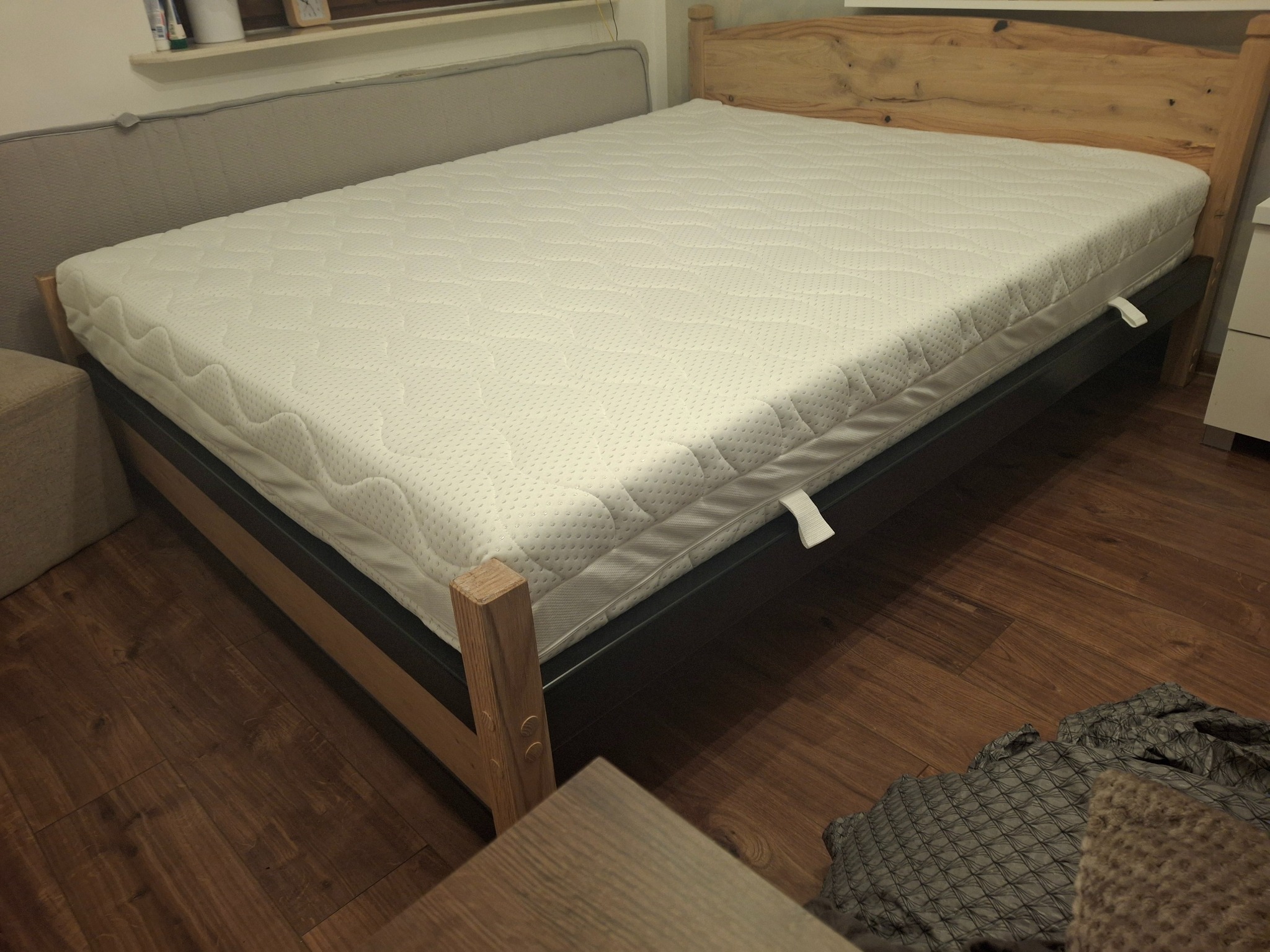 Drewniane łóżko z materacem, z widocznym stelażem i zagłówkiem. Na podłodze panele. Częściowo widoczna szafka nocna i kanapa. Stonowana kolorystyka.