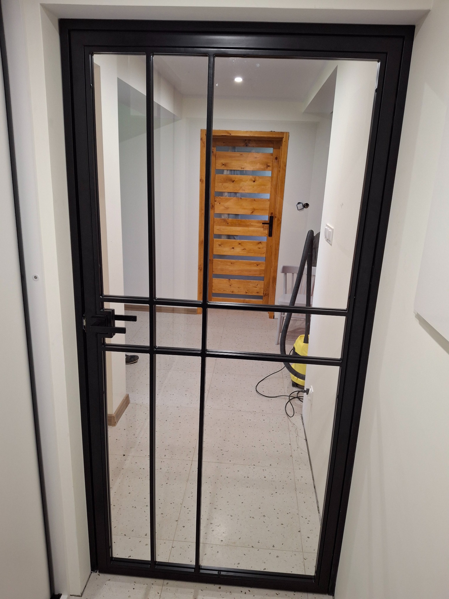 Czarna, metalowa rama z szybami, tworząca drzwi wewnętrzne w nowoczesnym stylu, widok z bliska na korytarz z drewnianymi drzwiami w tle.