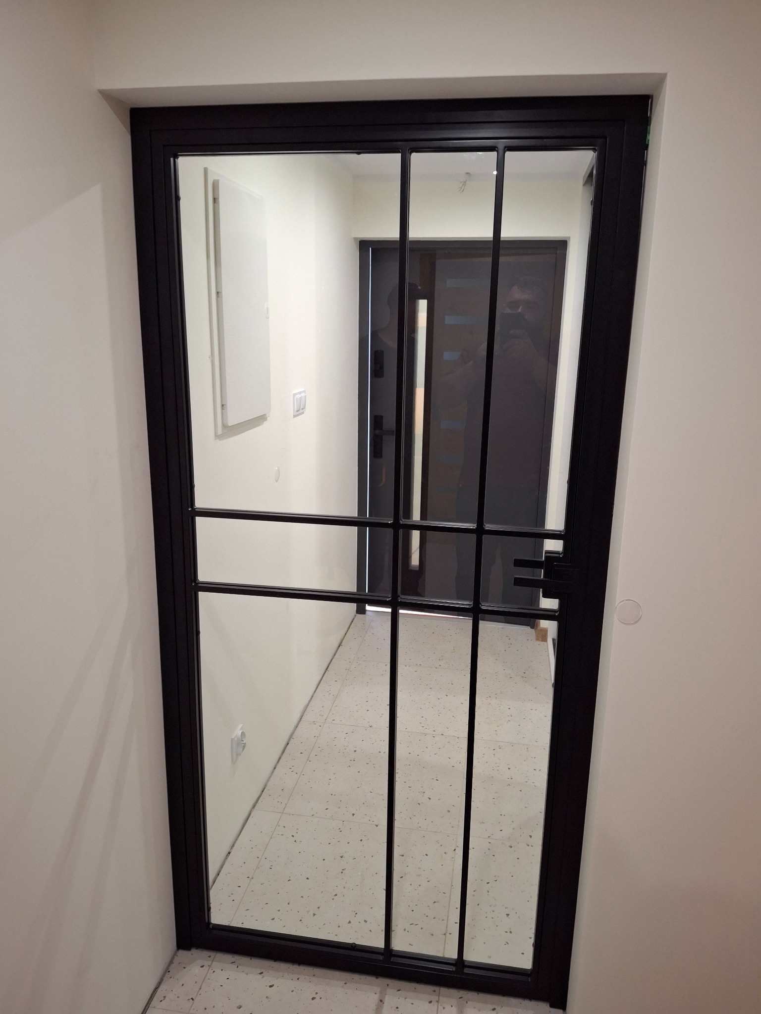 Czarna, metalowa konstrukcja z szybą, tworząca drzwi wewnętrzne z podziałami, widok na korytarz z płytkami lastryko.