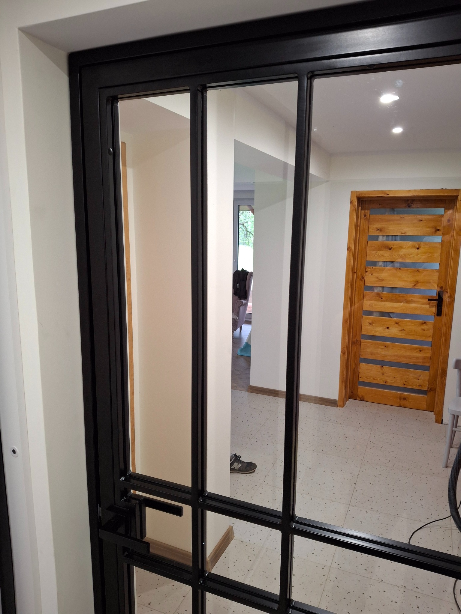 Nowoczesne drzwi wewnętrzne z czarnymi metalowymi szprosami i szybami, widok na jasne wnętrze z drewnianymi drzwiami i podłogą w terazzo.