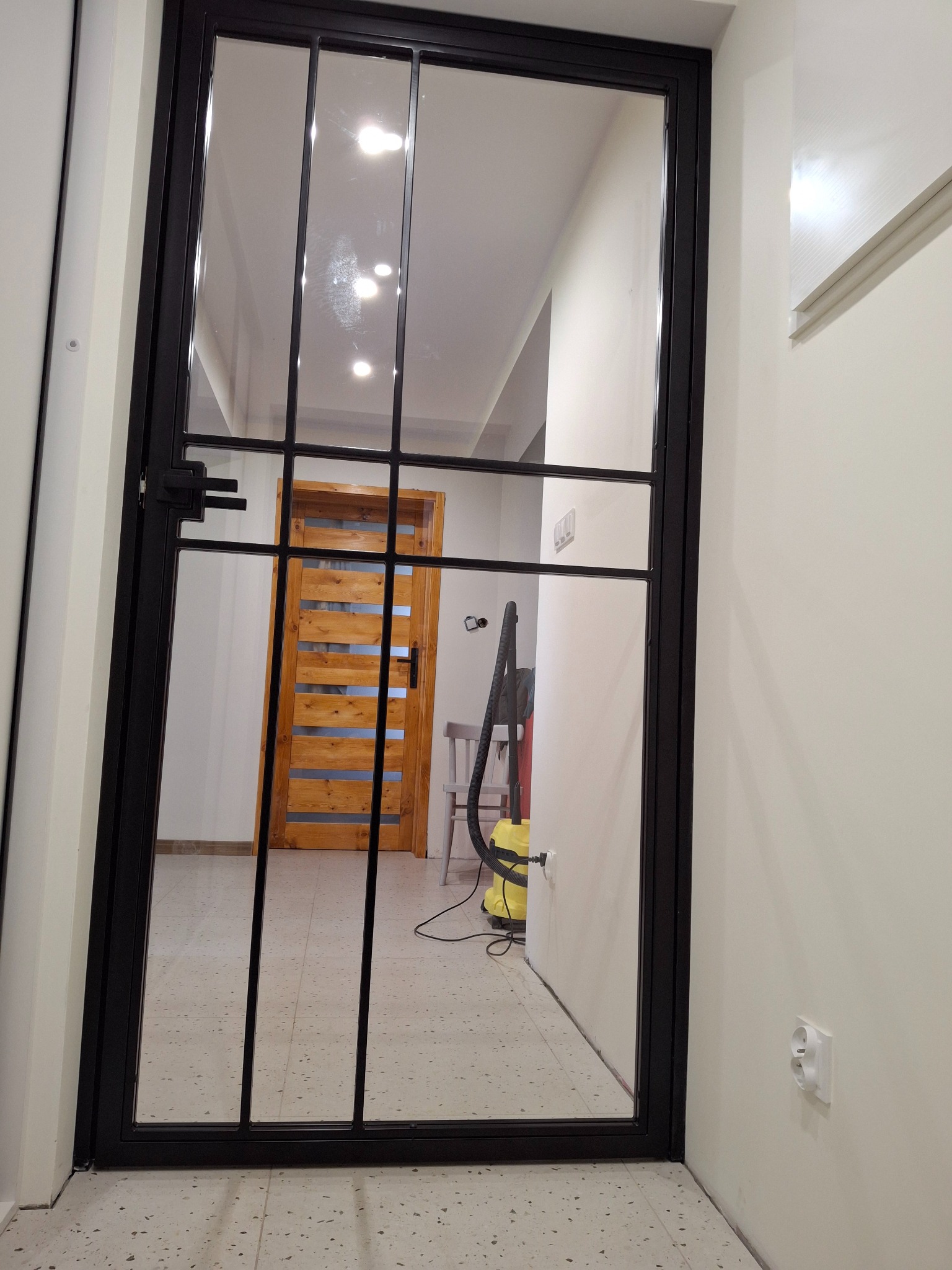 Czarna, metalowa ścianka działowa z szybami w korytarzu. Widoczna drewniana, ażurowa  ściana w tle. Minimalistyczny design,  jasne wnętrze.