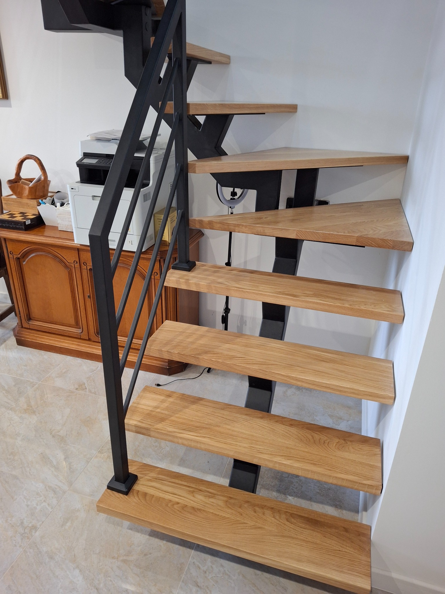 Nowoczesne schody z drewnianymi stopniami i czarną metalową balustradą, widok z dołu. W tle biurko i drukarka.