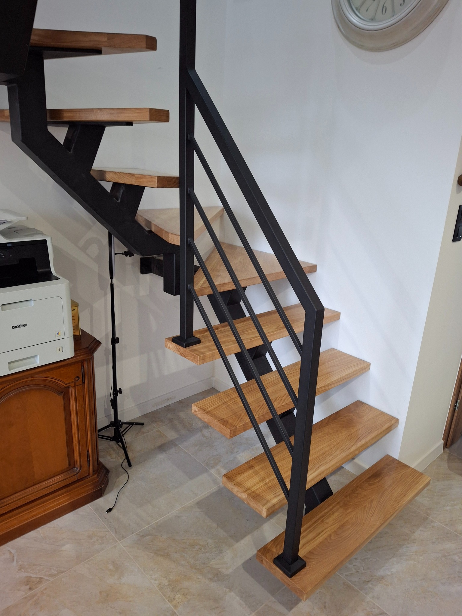 Nowoczesne schody z drewnianymi stopniami i czarną, minimalistyczną balustradą. Widok z dołu, ukazujący konstrukcję i detale wykończenia. Jasne wnętrze.