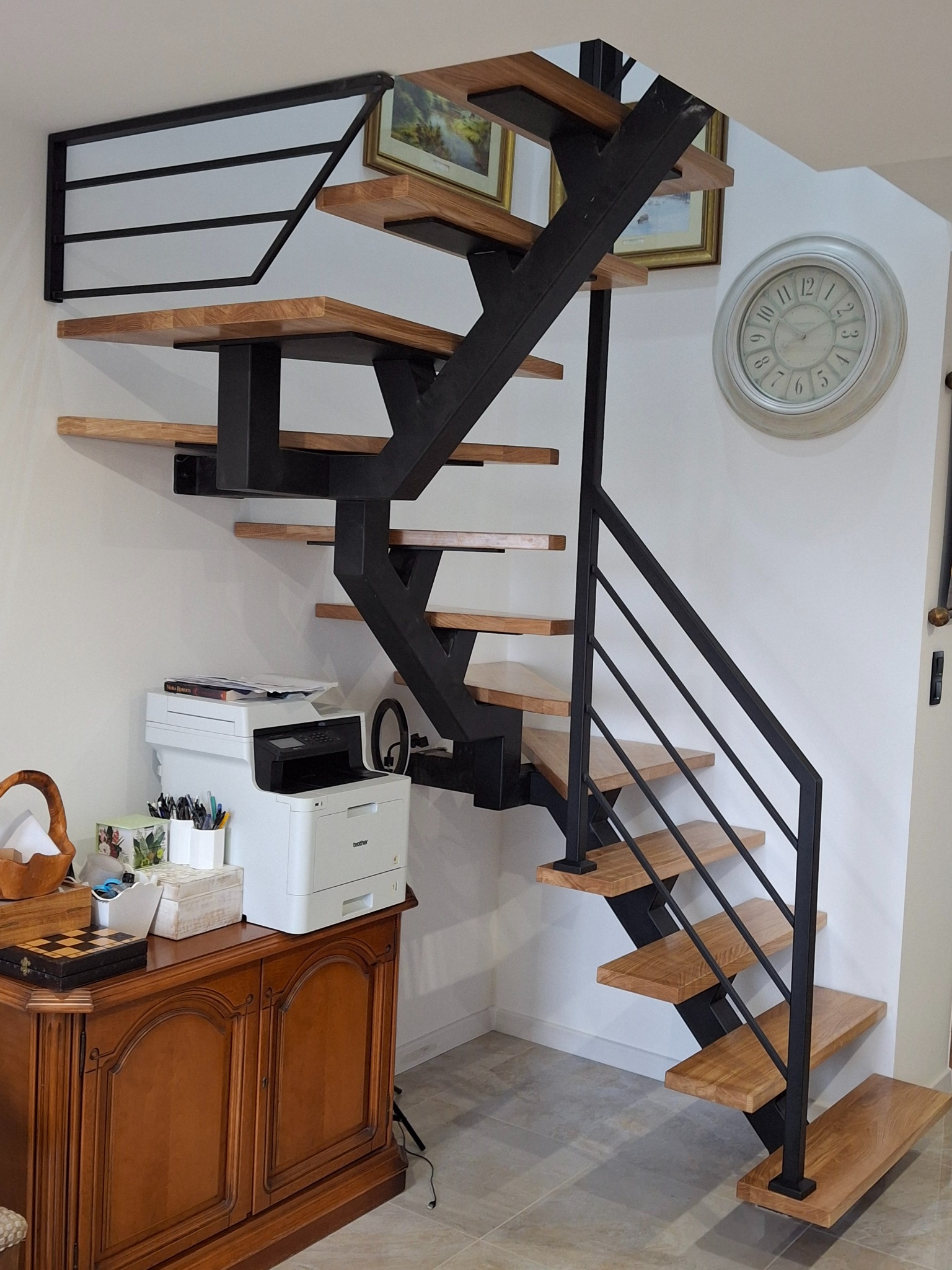Nowoczesne schody z drewnianymi stopniami i czarną metalową konstrukcją, zamontowane w jasnym wnętrzu. Widoczny fragment komody i drukarki.