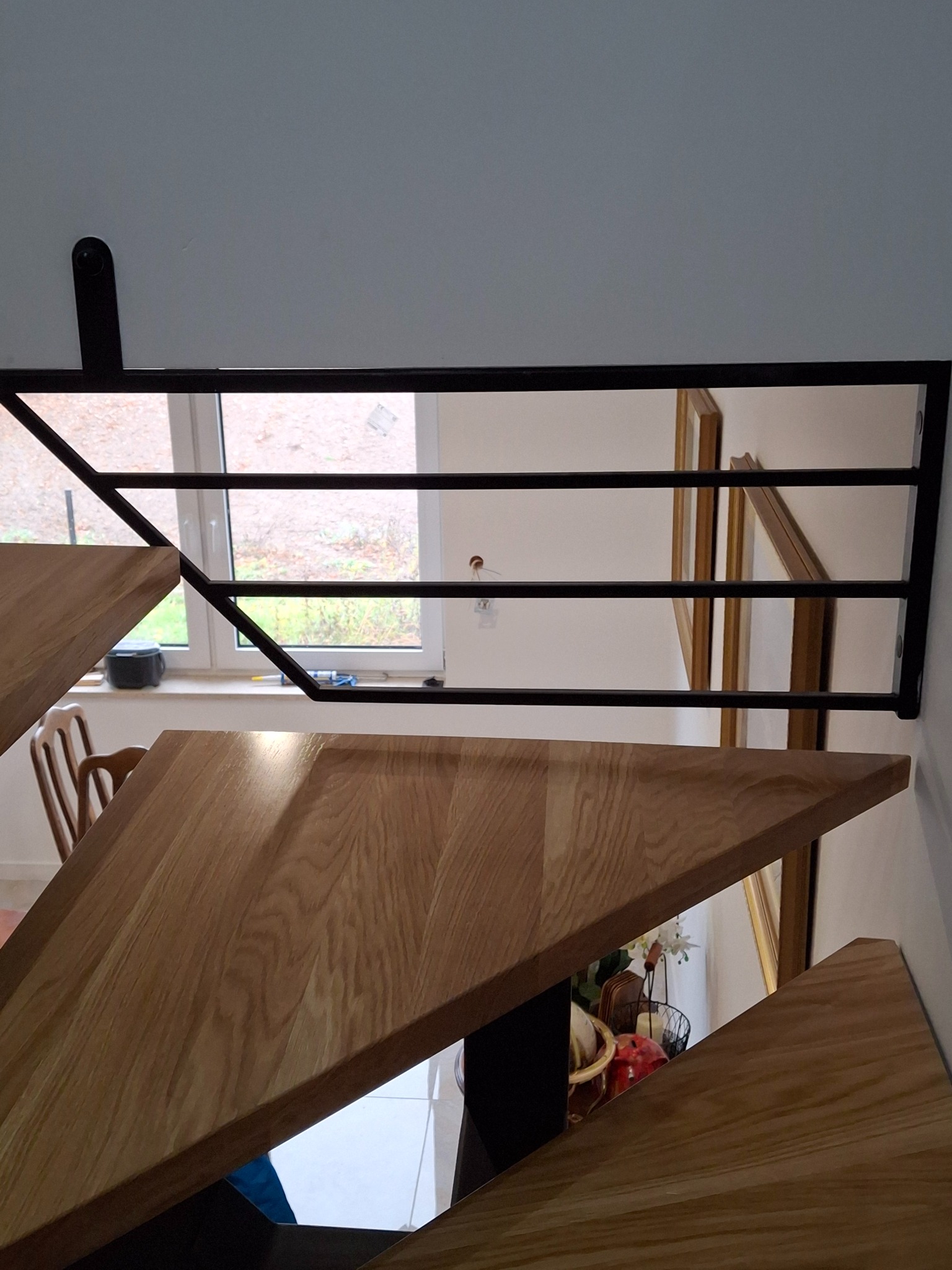 Nowoczesne schody drewniane z metalową balustradą, widok z góry. Stopnie o trójkątnym kształcie, jasne drewno. W tle okno i dekoracje.