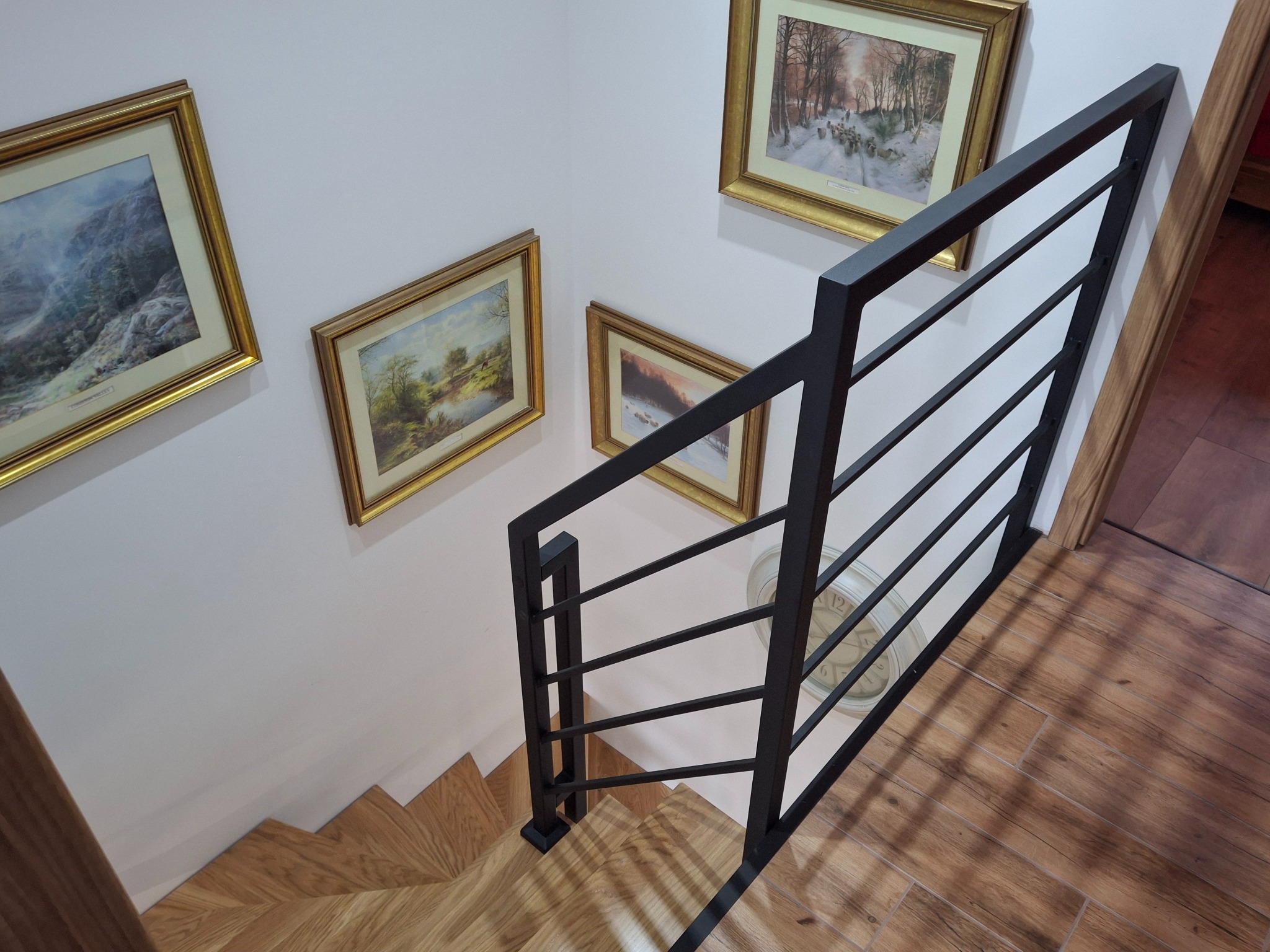 Nowoczesne schody z drewnianymi stopniami i czarną, minimalistyczną balustradą. Na ścianie wiszą obrazy w złotych ramach. Widok z góry, podkreślający geometrię.