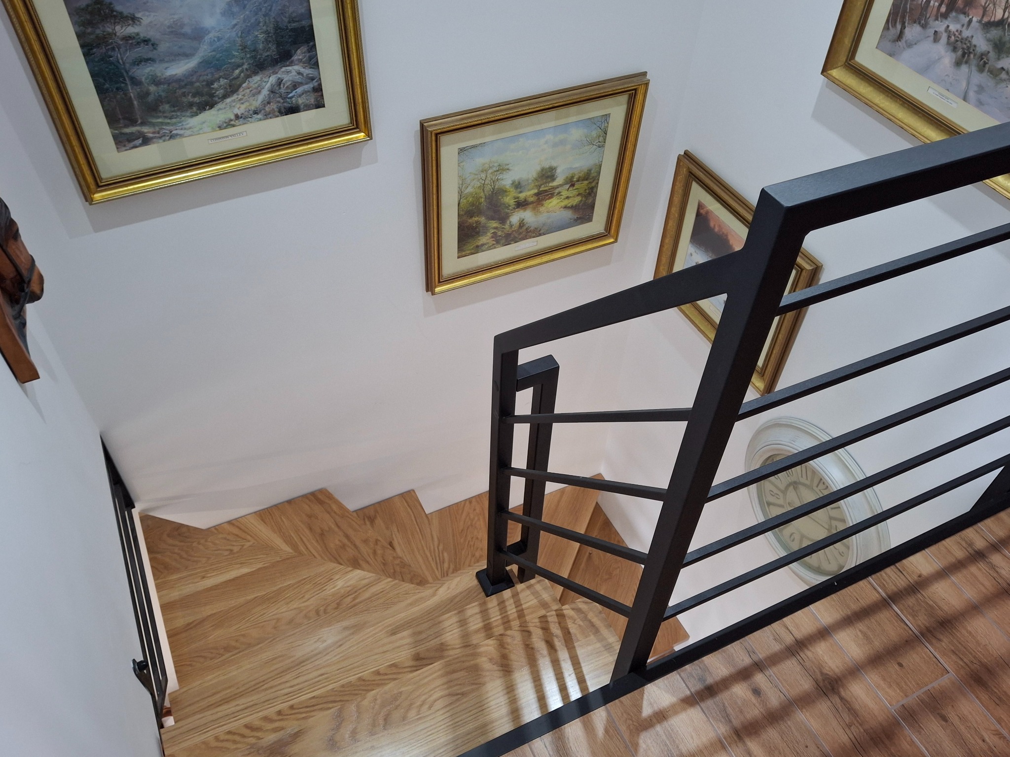 Nowoczesne schody z drewnianymi stopniami i czarną, minimalistyczną balustradą. Ujęcie z góry, widoczne obrazy na ścianie i zegar.