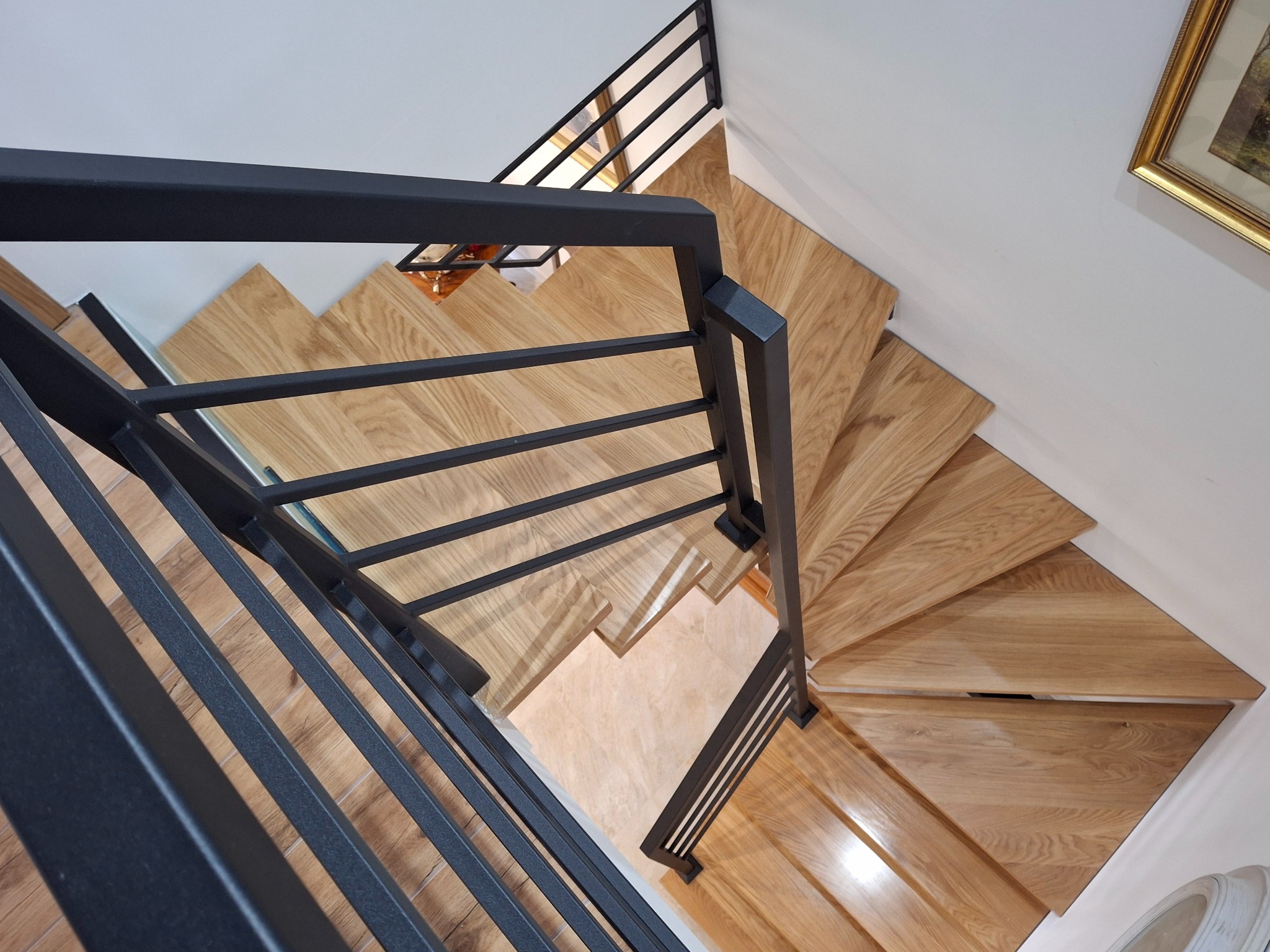 Spiralne schody z jasnego drewna z czarną, metalową balustradą, widziane z góry. U góry fragment ściany z obrazem w złotej ramie. Nowoczesny design.
