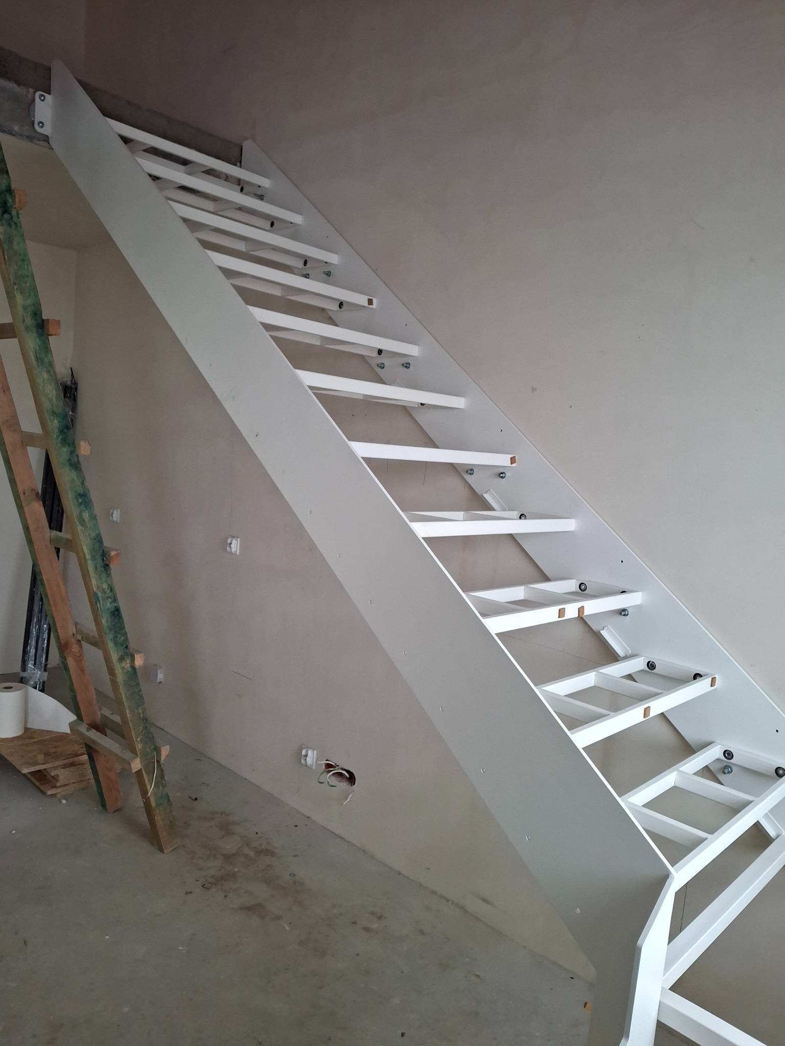 Białe schody z drewnianymi stopniami w trakcie montażu, widoczne elementy konstrukcyjne i drabina oparta o ścianę. Surowy stan wykończenia wnętrza.