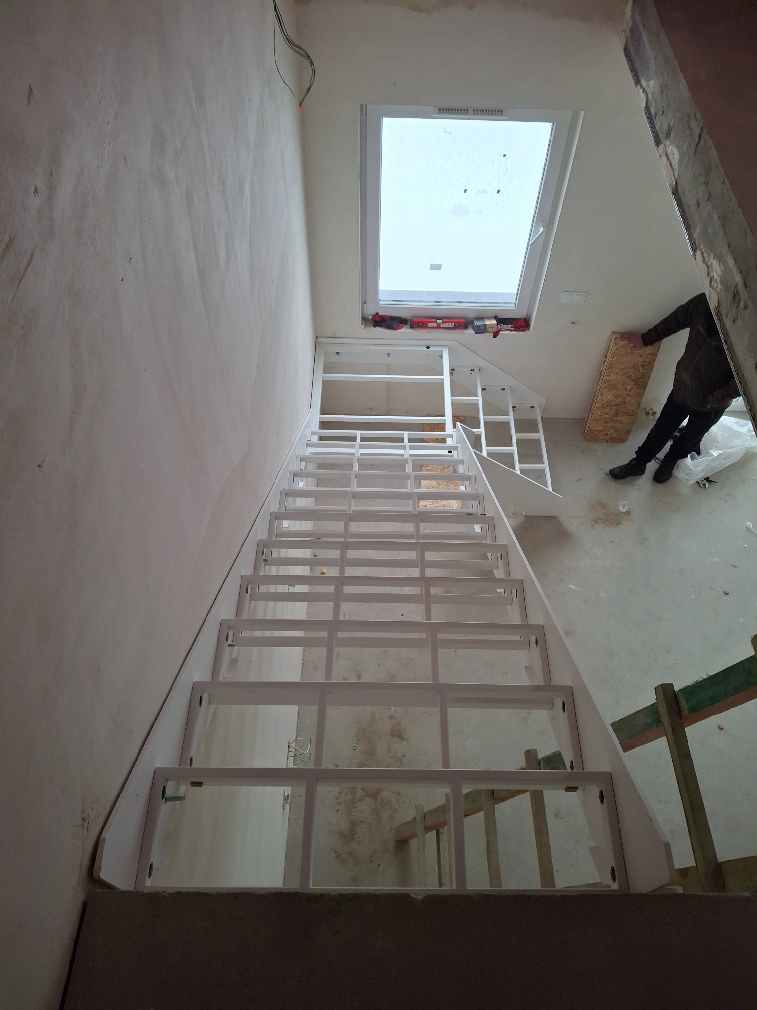 Białe, surowe schody z metalową konstrukcją w trakcie montażu w jasnym wnętrzu z oknem dachowym. Widoczne narzędzia i fragment osoby przy schodach.