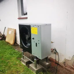 Zewnętrzna jednostka szaro-czarnej pompy ciepła zamontowana na metalowej konstrukcji wspartej na betonowych bloczkach, umieszczona na trawniku przy białej ścianie budynku z widocznym oknem...