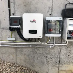 Instalacja elektryczna z białym inwerterem Afore na surowej betonowej ścianie, widoczne kable w peszlach i skrzynki rozdzielcze, u dołu kamieniste podłoże.