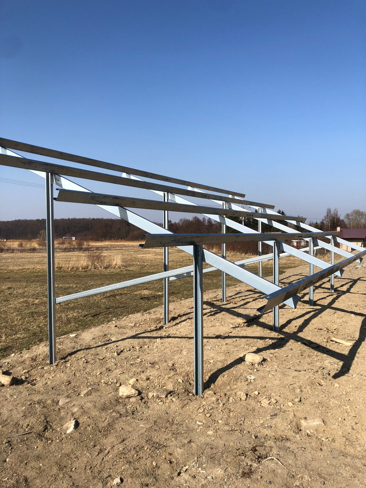 Metalowa konstrukcja wsporcza pod panele słoneczne w trakcie montażu, widoczny krajobraz polny z zabudowaniami w tle, błękitne niebo.