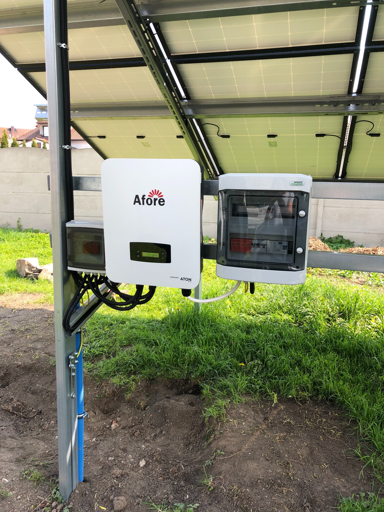 Inwerter Afore i skrzynka zabezpieczeń AC zamontowane pod panelami słonecznymi, widoczne kable w peszlu i trawa w tle.