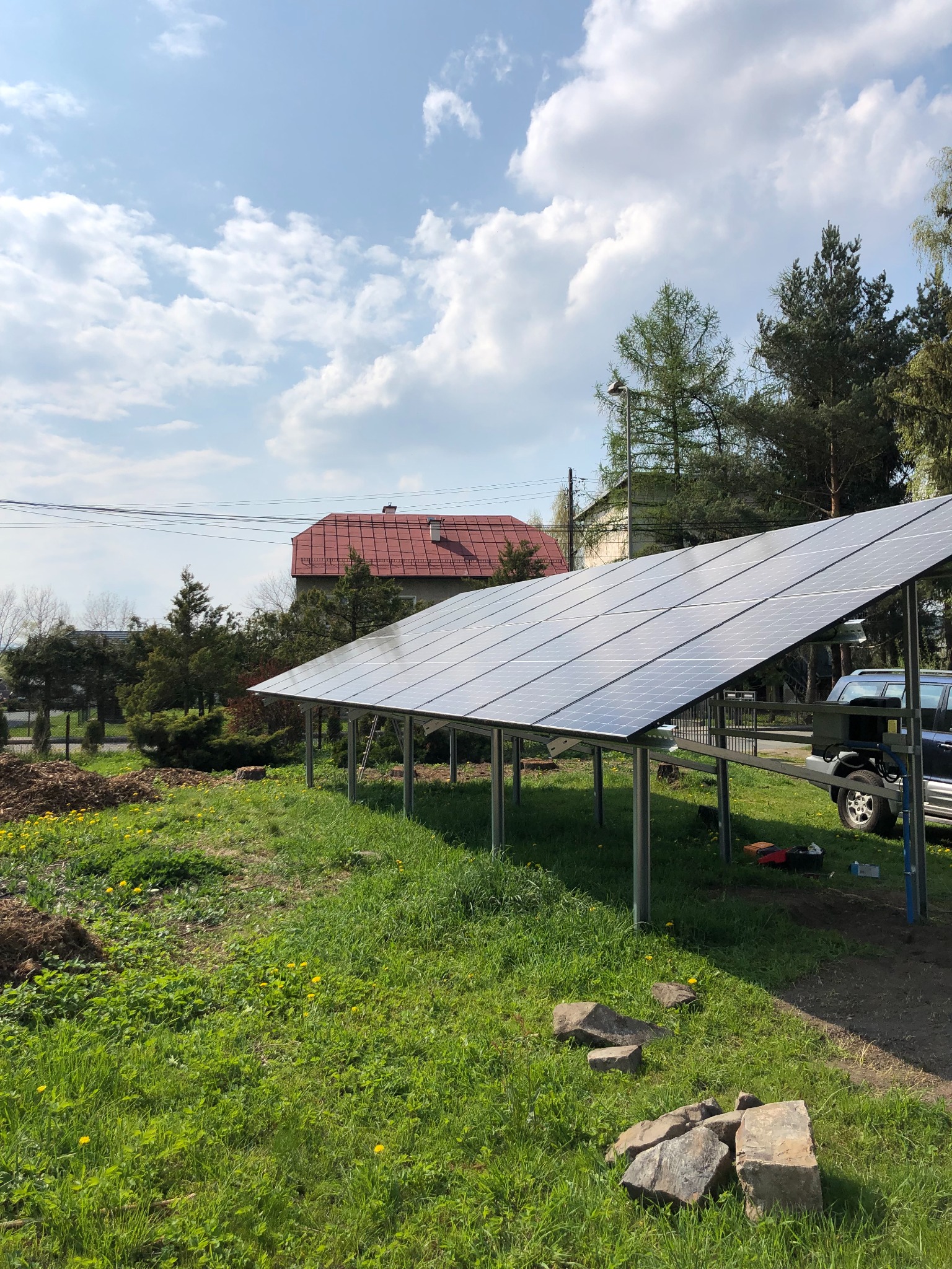 Instalacja paneli słonecznych na metalowej konstrukcji w ogrodzie, z widocznym samochodem i narzędziami, dom z czerwoną dachówką w tle, słoneczny dzień.