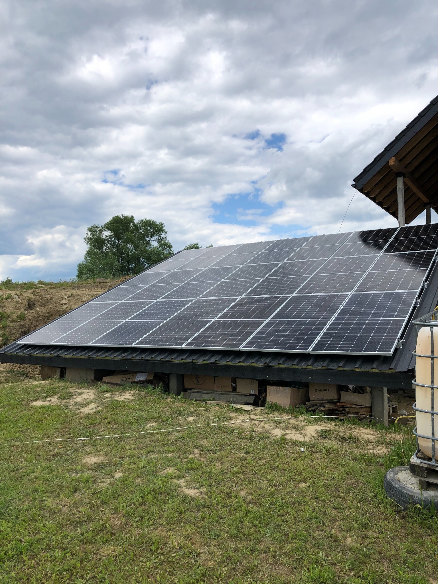 Instalacja fotowoltaiczna na dachu budynku gospodarczego z widocznymi kartonami i zbiornikiem na wodę w otoczeniu trawy i ziemi, pochmurne niebo w tle.