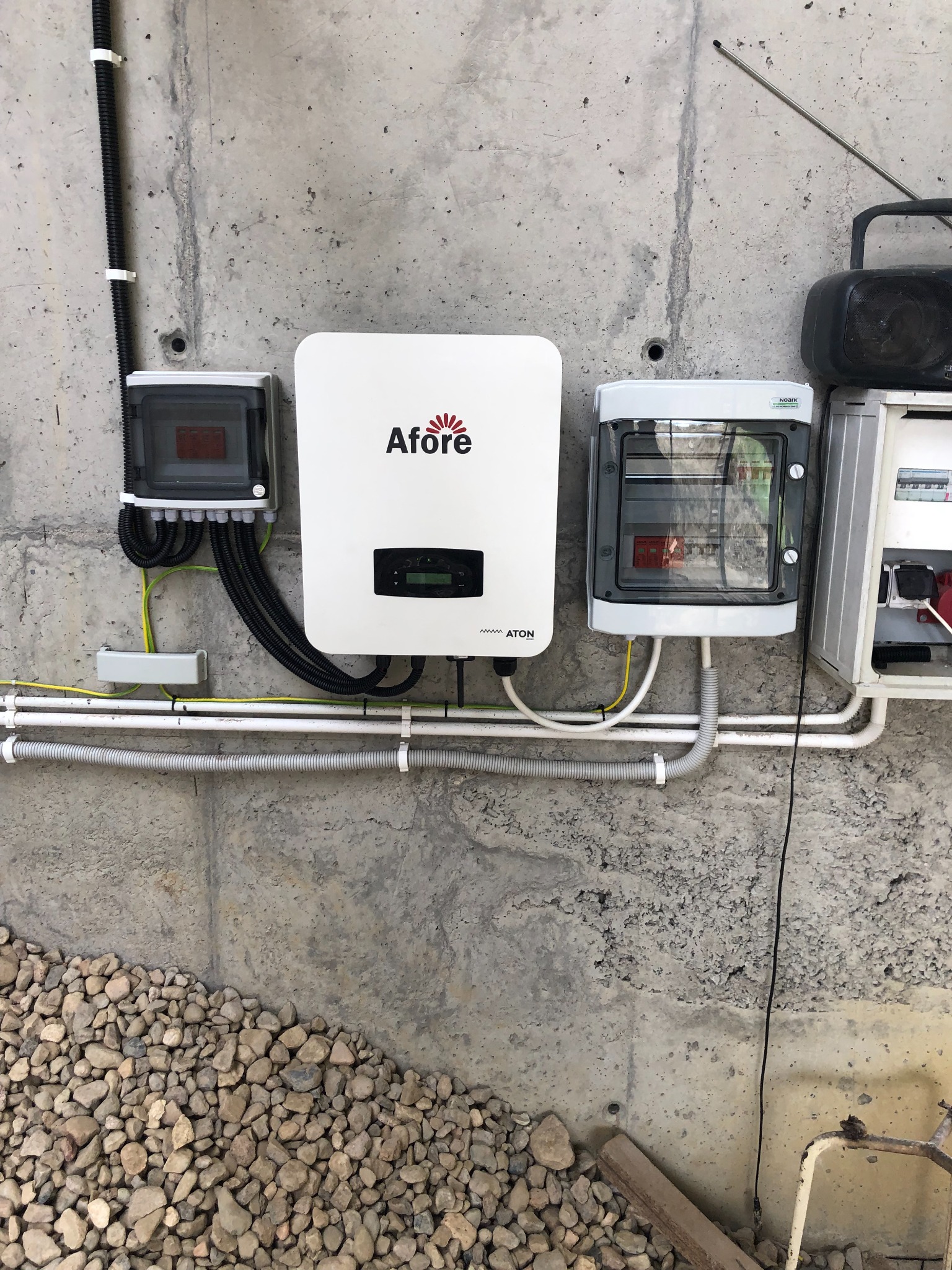 Instalacja elektryczna z białym inwerterem Afore na surowej betonowej ścianie, widoczne kable w peszlach i skrzynki rozdzielcze, u dołu kamieniste podłoże.