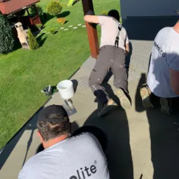 Montaż tarasu na zewnątrz: pracownicy w koszulkach z logo firmy układają szarą nawierzchnię wokół drewnianego słupa, w tle widoczny trawnik i plac zabaw.