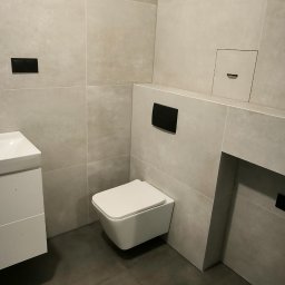 PWW Profesjonalne Wykończenia Wnętrz - Minimalistyczna łazienka z wiszącą toaletą, umywalką i szarymi płytkami imitującymi beton, podkreślona czarnymi akcentami w postaci przycisków spłukiwania.