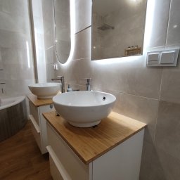 PWW Profesjonalne Wykończenia Wnętrz - Nowoczesna łazienka z dwoma umywalkami nablatowymi w kształcie misy, drewnianymi blatami i podświetlanymi lustrami owalnymi, wykończona płytkami w odcieniach beżu i szarości.