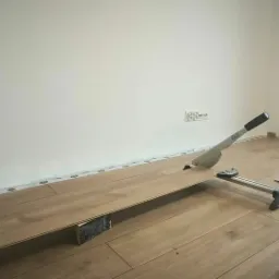 Przycinanie panelu laminowanego za pomocą gilotyny do paneli, widoczne listwy przypodłogowe, poziomica, miarka i młotek na podłodze w trakcie montażu.