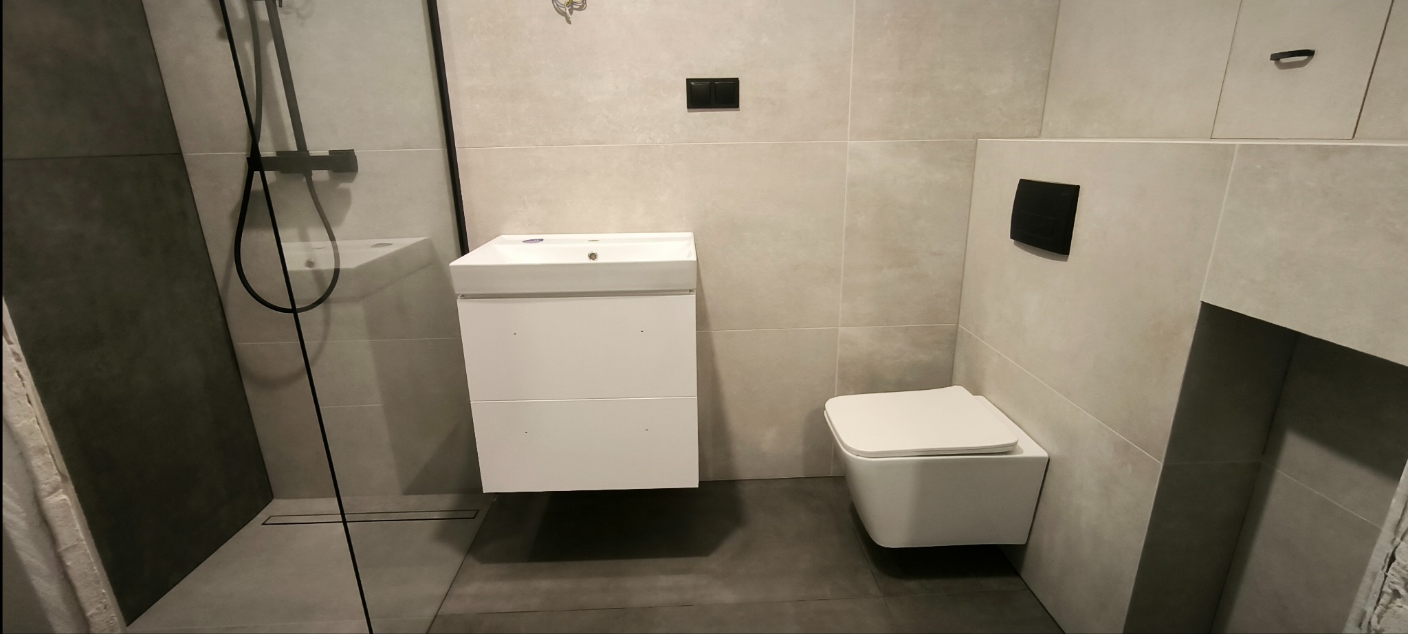 Nowoczesna łazienka z kabiną prysznicową typu walk-in, podwieszaną szafką z umywalką i toaletą w kolorze białym, wykończona płytkami w odcieniach szarości.